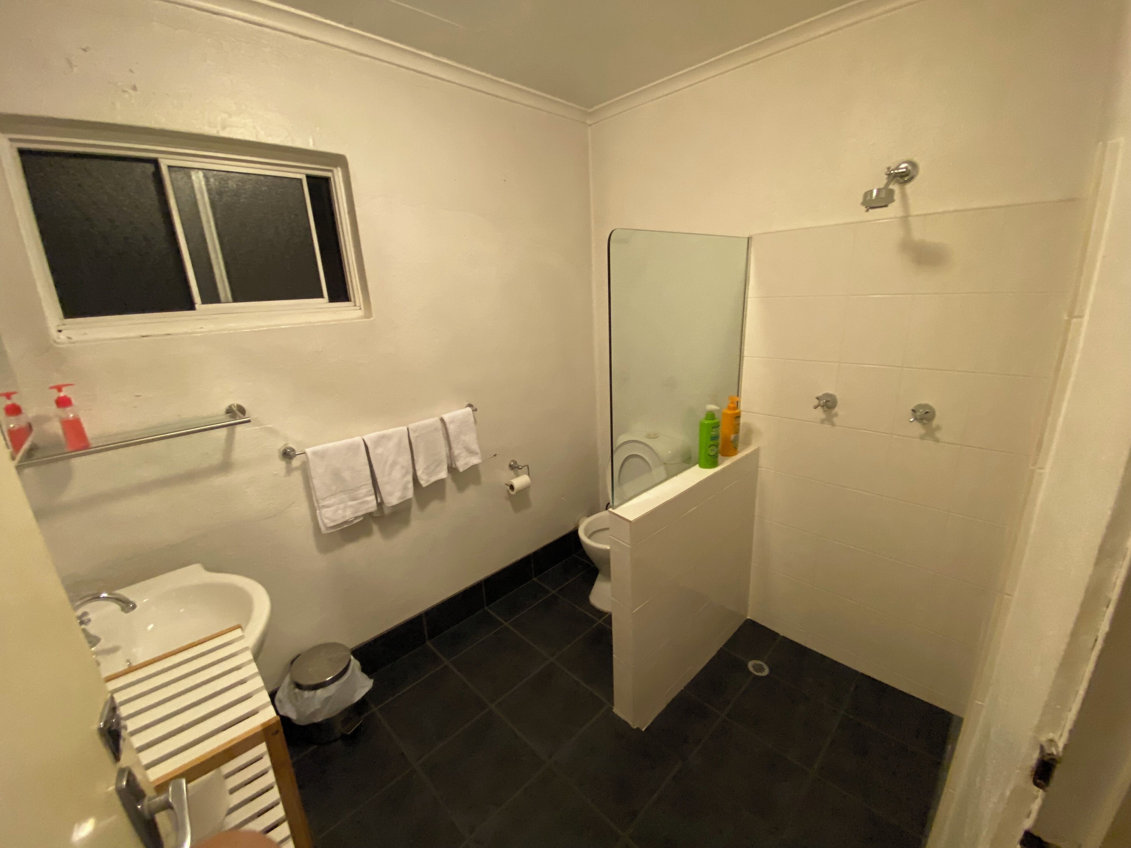 Un baño pequeño con inodoro, lavamanos y espejo. Hay una ventana sobre el inodoro y un perchero con toallas.