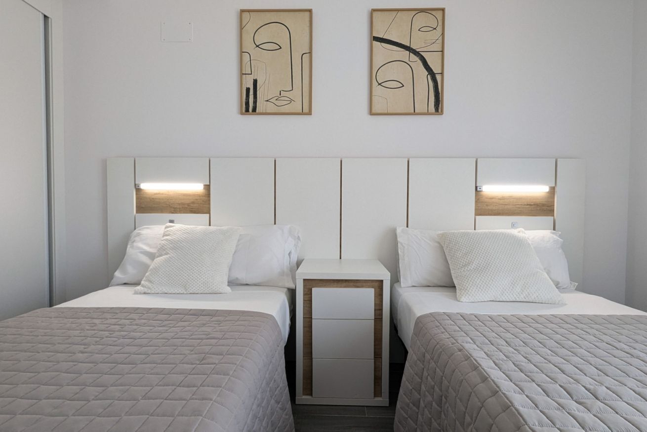 A modern hotel room with two single beds, a bedside table, and a headboard with integrated lighting.