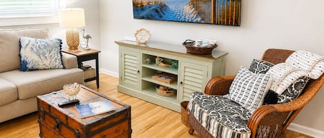 Una sala de estar con un sofá, una silla de mimbre, un cofre de madera, un televisor montado en la pared y un mueble bajo con cajones.