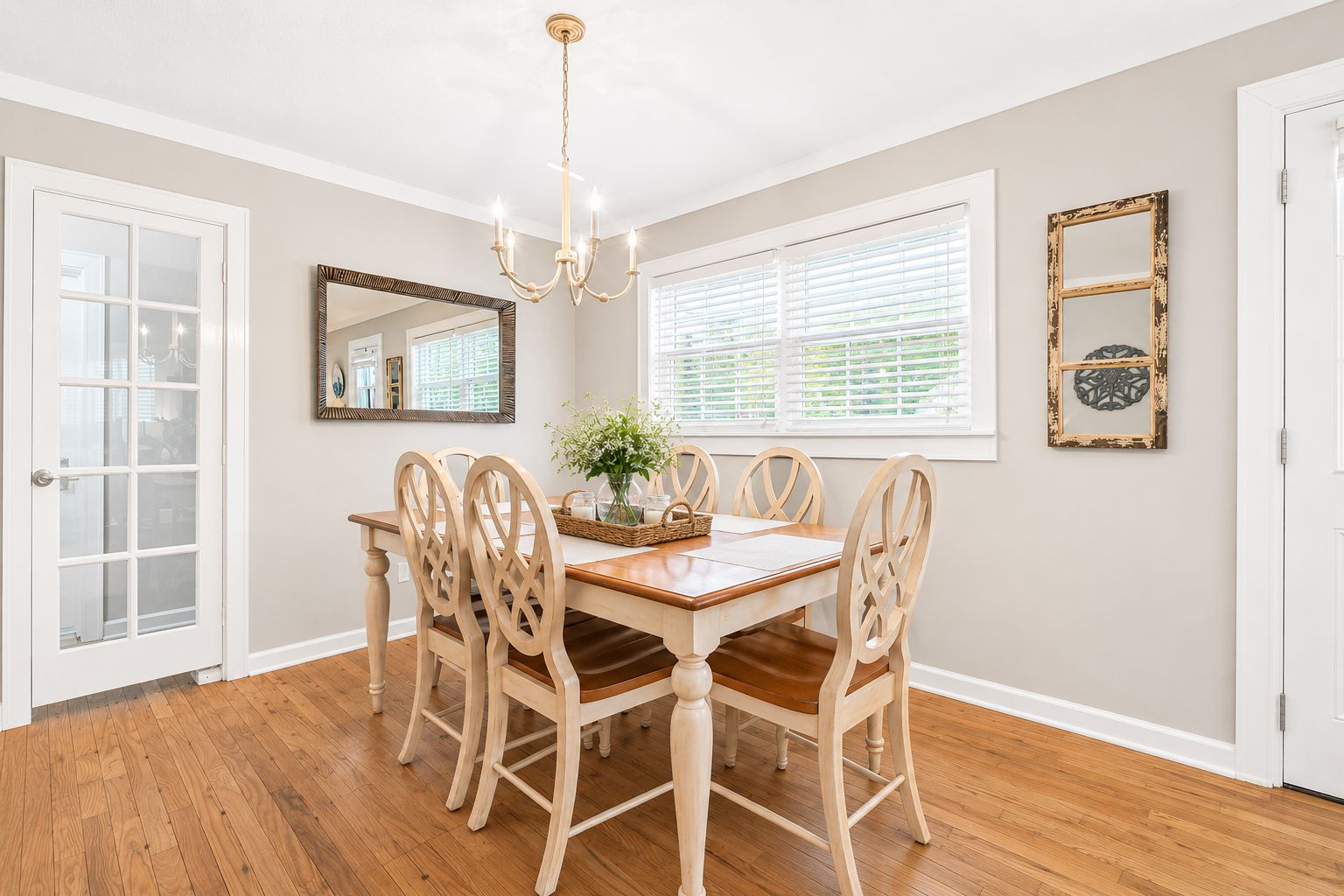 Une salle à manger avec une table et des chaises en bois, un lustre, un miroir et une fenêtre avec des stores.