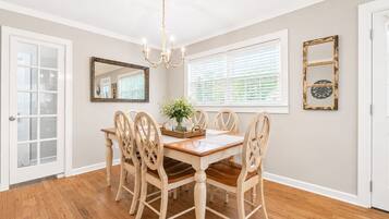 Une salle à manger avec une table et des chaises en bois, un lustre, un miroir et une fenêtre avec des stores.