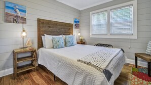 Une chambre à coucher avec une tête de lit en bois, un grand lit avec une literie blanche, deux tables de chevet avec des lampes et une fenêtre avec des stores.