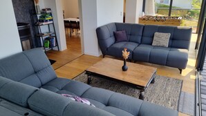 Een moderne woonkamer met een grijze bank, een salontafel en een eethoek, zichtbaar door een open deur.