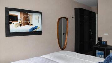 A hotel room with a bed, a TV mounted on the wall displaying a room view, a mirror, and a wardrobe.