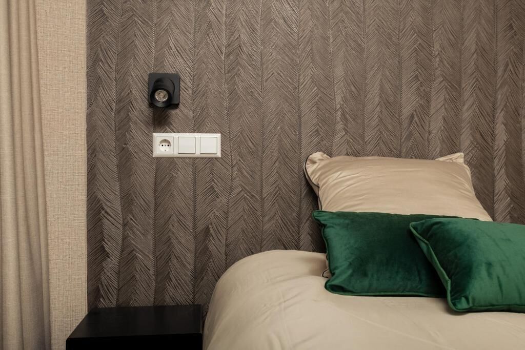 Una cama bien hecha con un cojín verde y uno beige, una mesita de noche negra, una lámpara fijada a la pared y un enchufe.