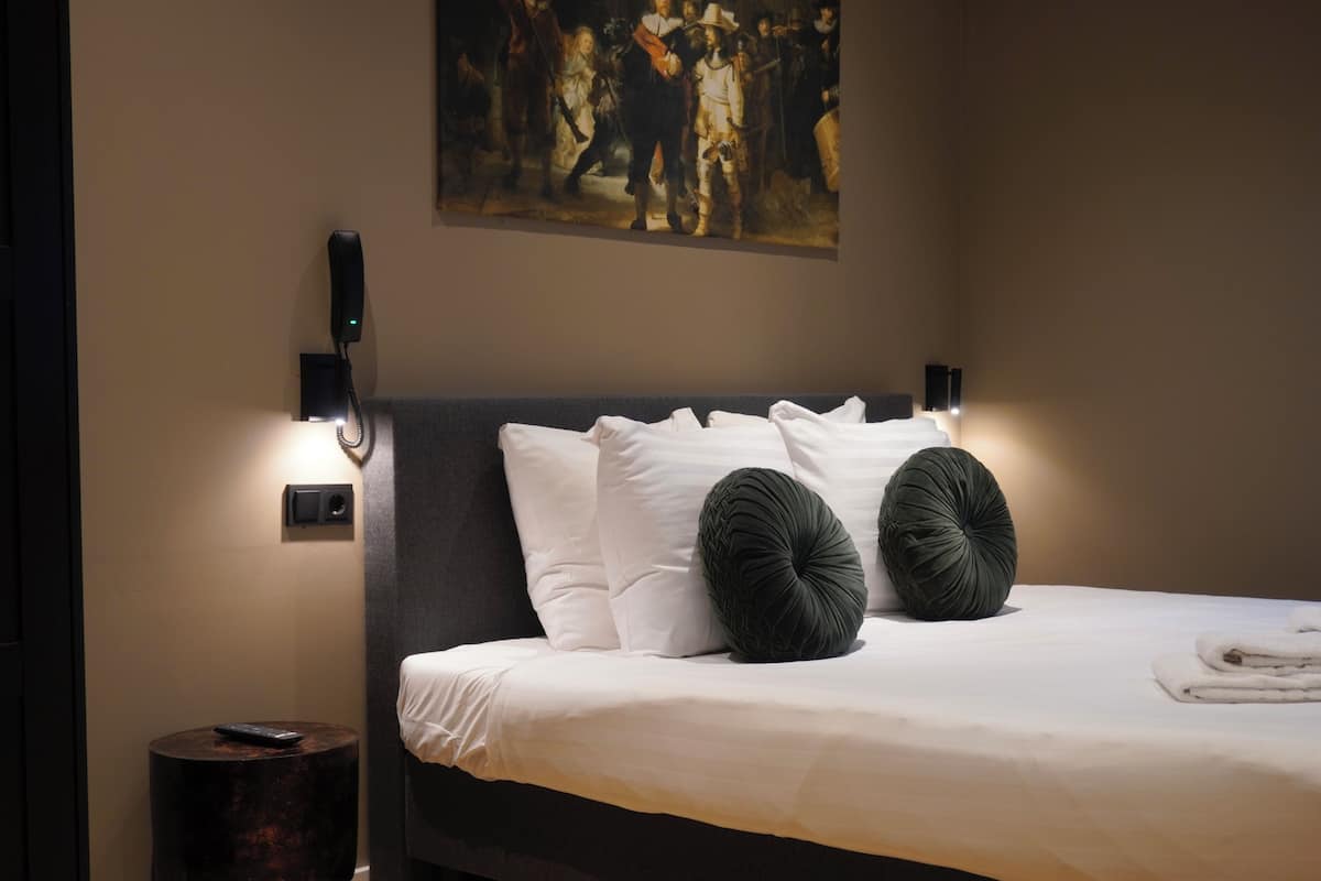 A neatly made bed with white linens and two dark green pillows, a wall-mounted painting, and a bedside table with a remote control.