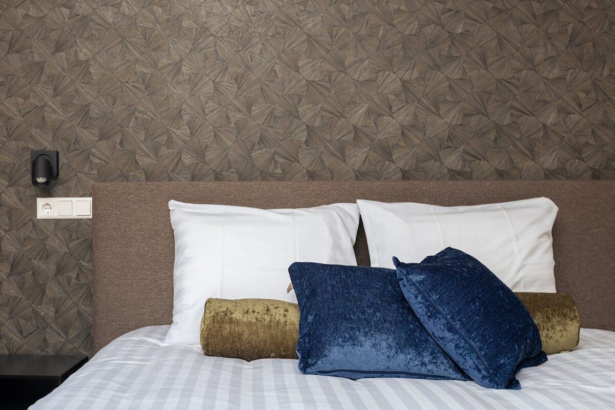 A neatly made bed with white and blue pillows, a textured wall, and a black nightstand with a wall socket.
