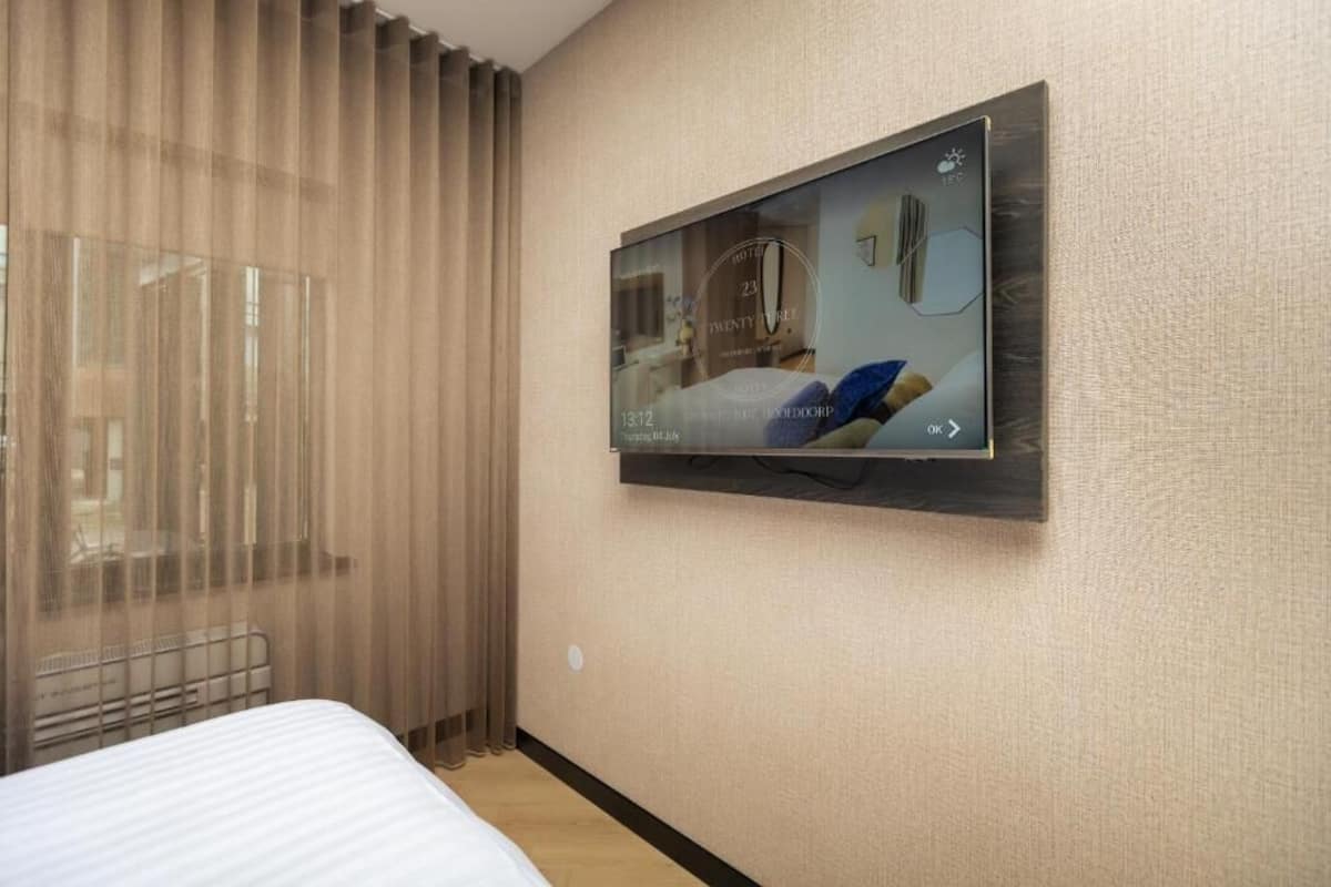 A hotel room with a bed, a TV mounted on the wall, and a window with sheer curtains.