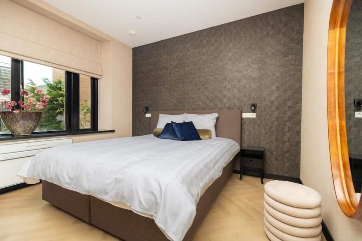 A modern bedroom with a large bed, a window with flowers, and a textured wall.