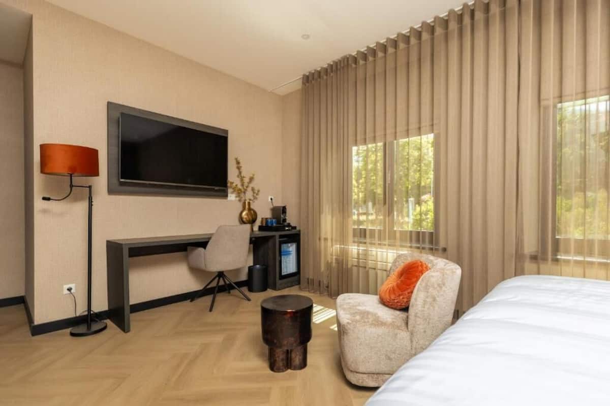 A modern hotel room with a bed, a desk with a TV, a chair, and a large window with curtains.