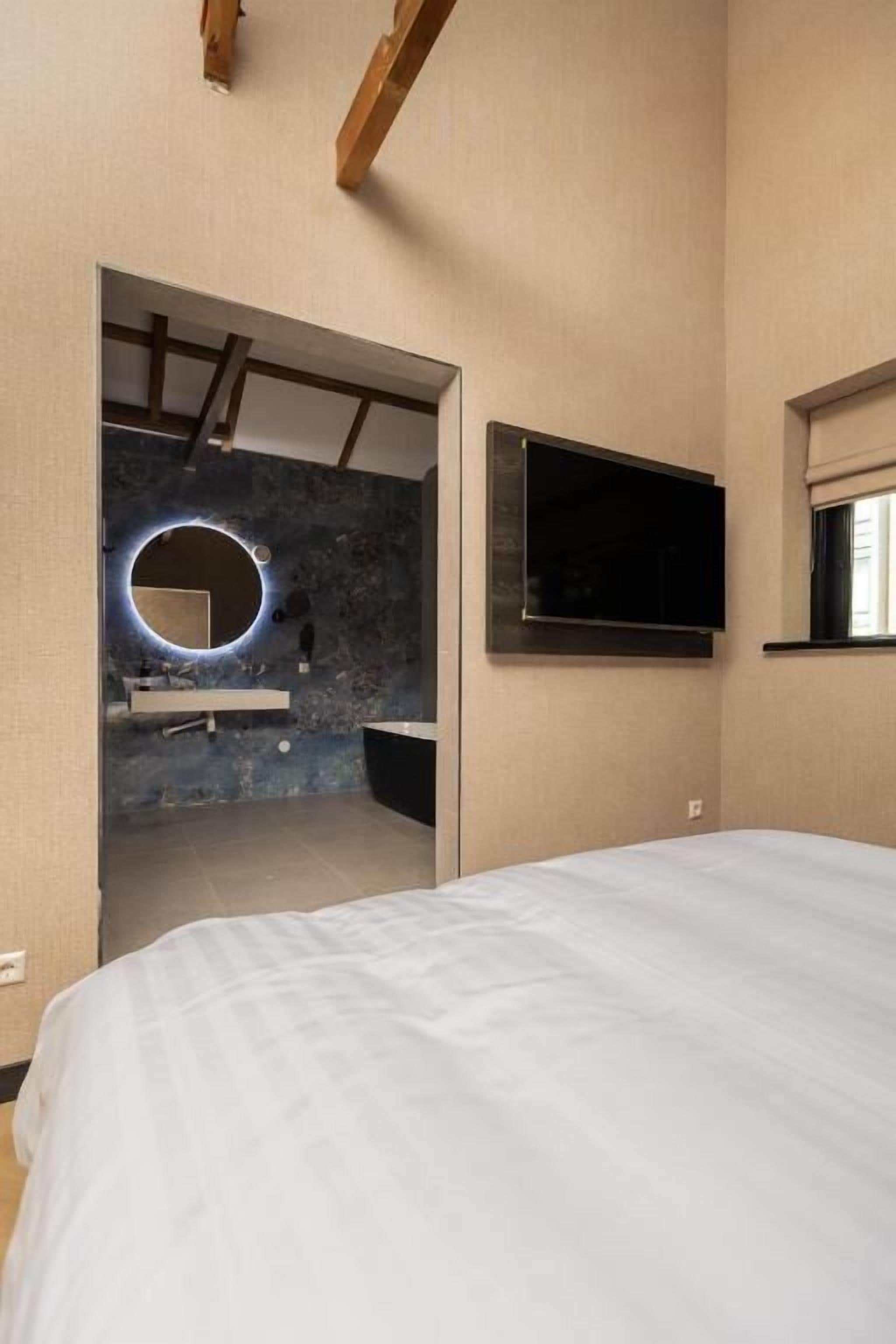 Une chambre à coucher avec un lit, un téléviseur fixé au mur et une salle de bain visible à travers une porte ouverte.