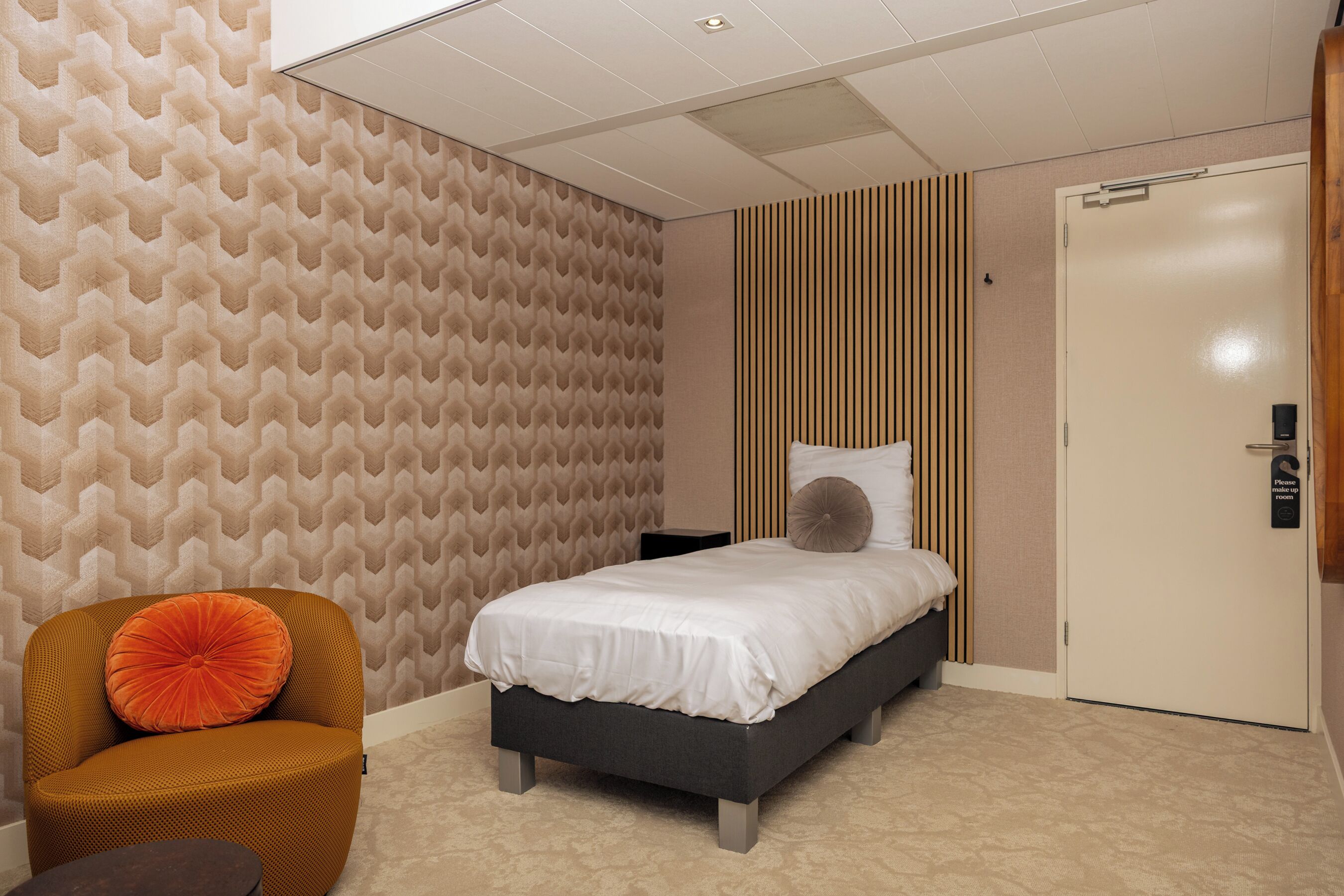 A modern bedroom with a patterned wallpaper, a bed with white bedding, a wooden chair, and a bedside table.