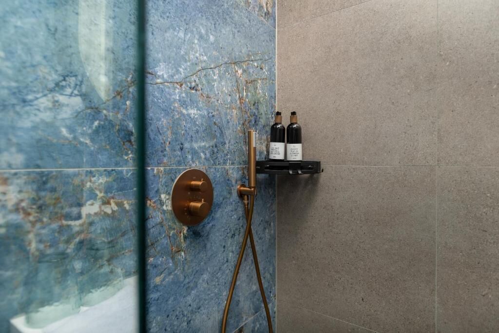 A bathroom with a blue marble wall, a gold showerhead, and two bottles on a shelf.