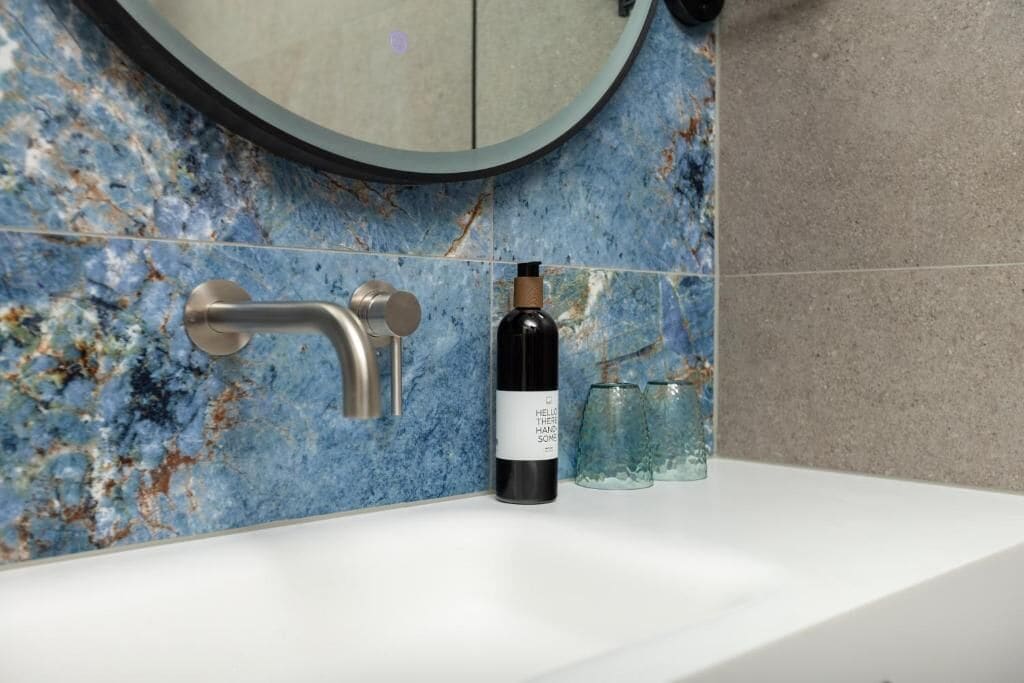 Un baño con lavamanos blanco, salpicadero de mármol azul, espejo redondo y una botella de jabón de manos.
