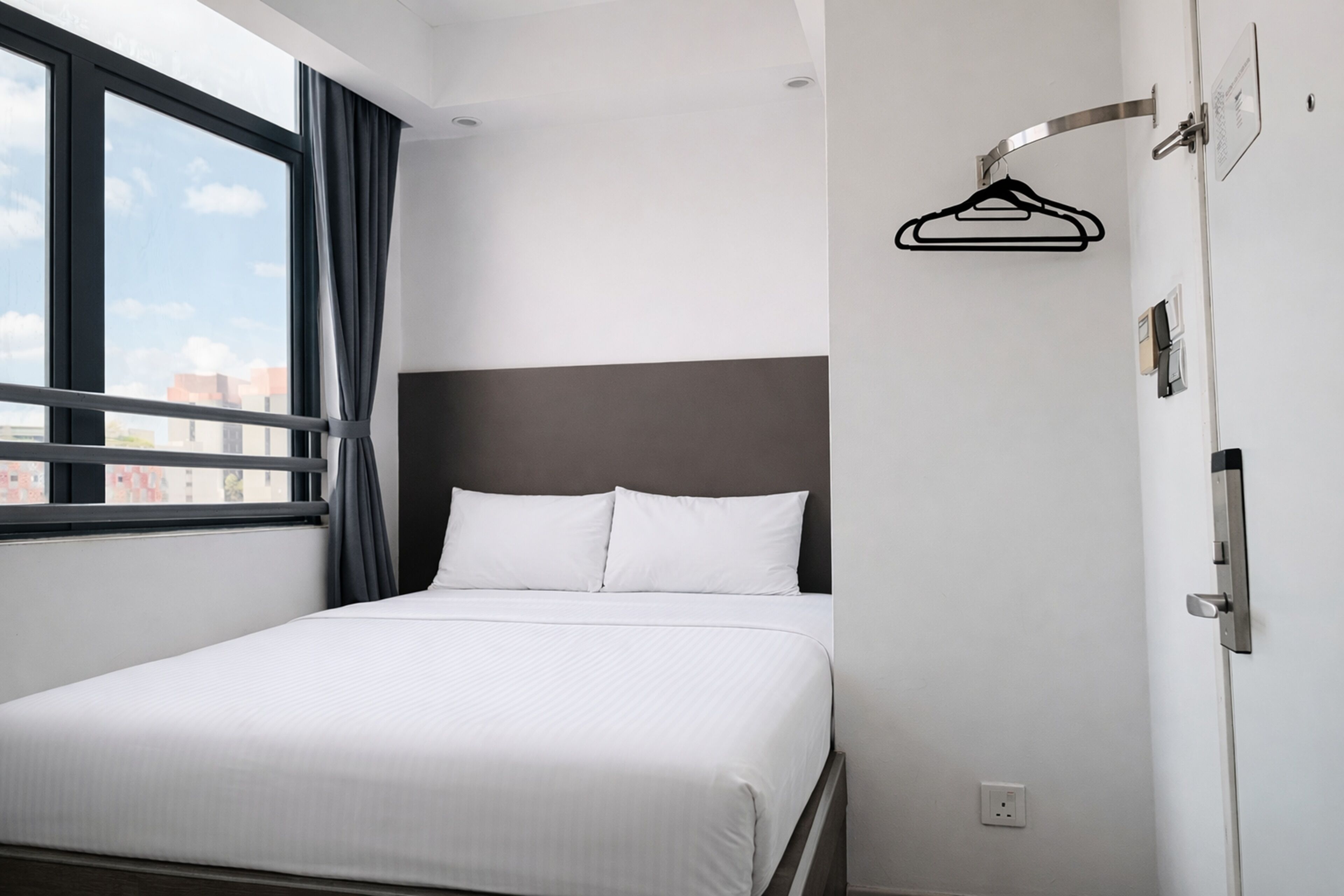Une chambre d’hôtel avec un lit, une fenêtre avec des rideaux, un porte-manteau fixé au mur et une porte avec une poignée.
