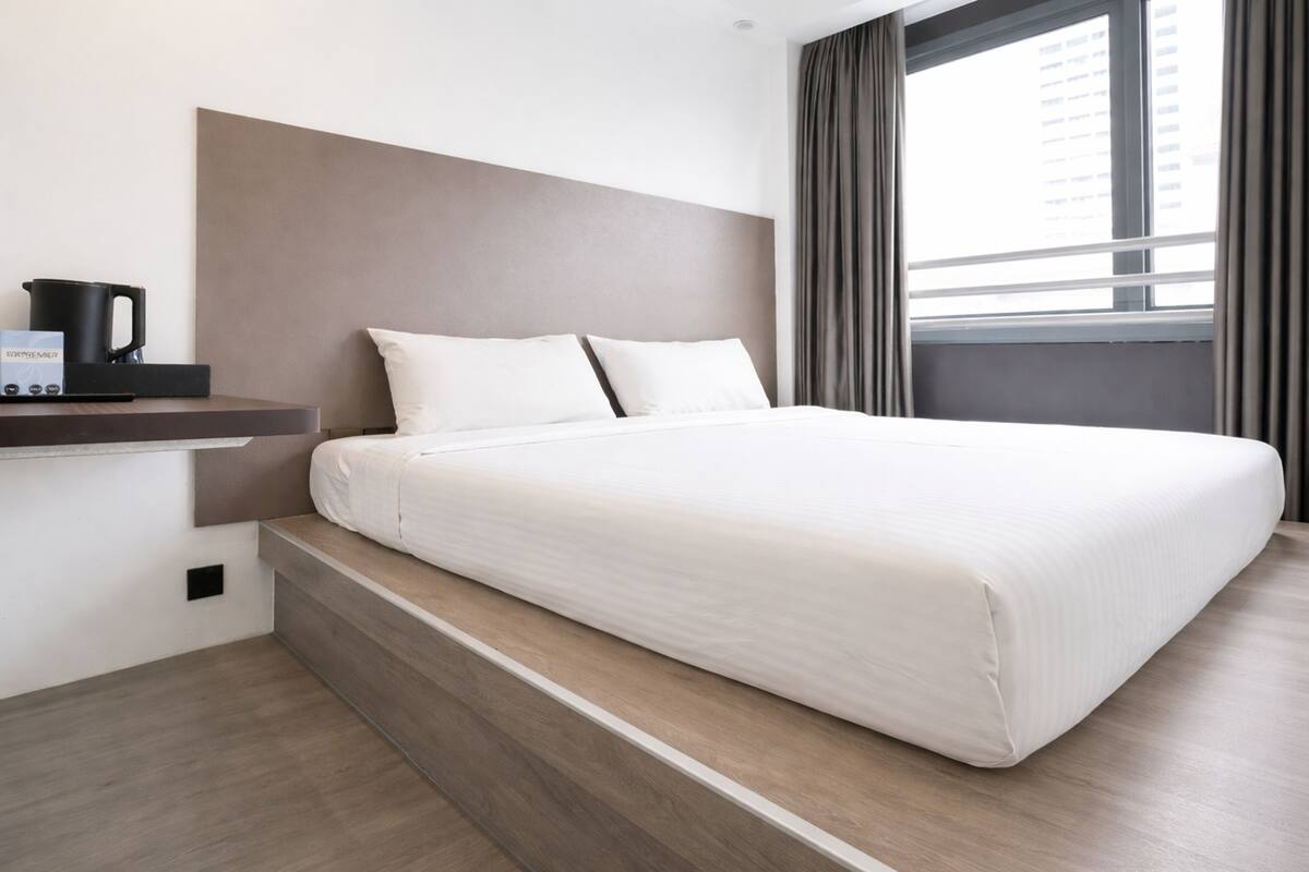 A modern hotel room with a large bed, a bedside table with a coffee maker, and a view of the city through a window.
