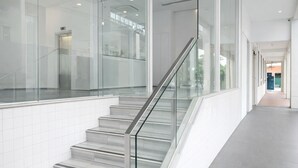 Un intérieur moderne avec un escalier à garde-corps en verre, des murs recouverts de carreaux blancs et un long couloir.