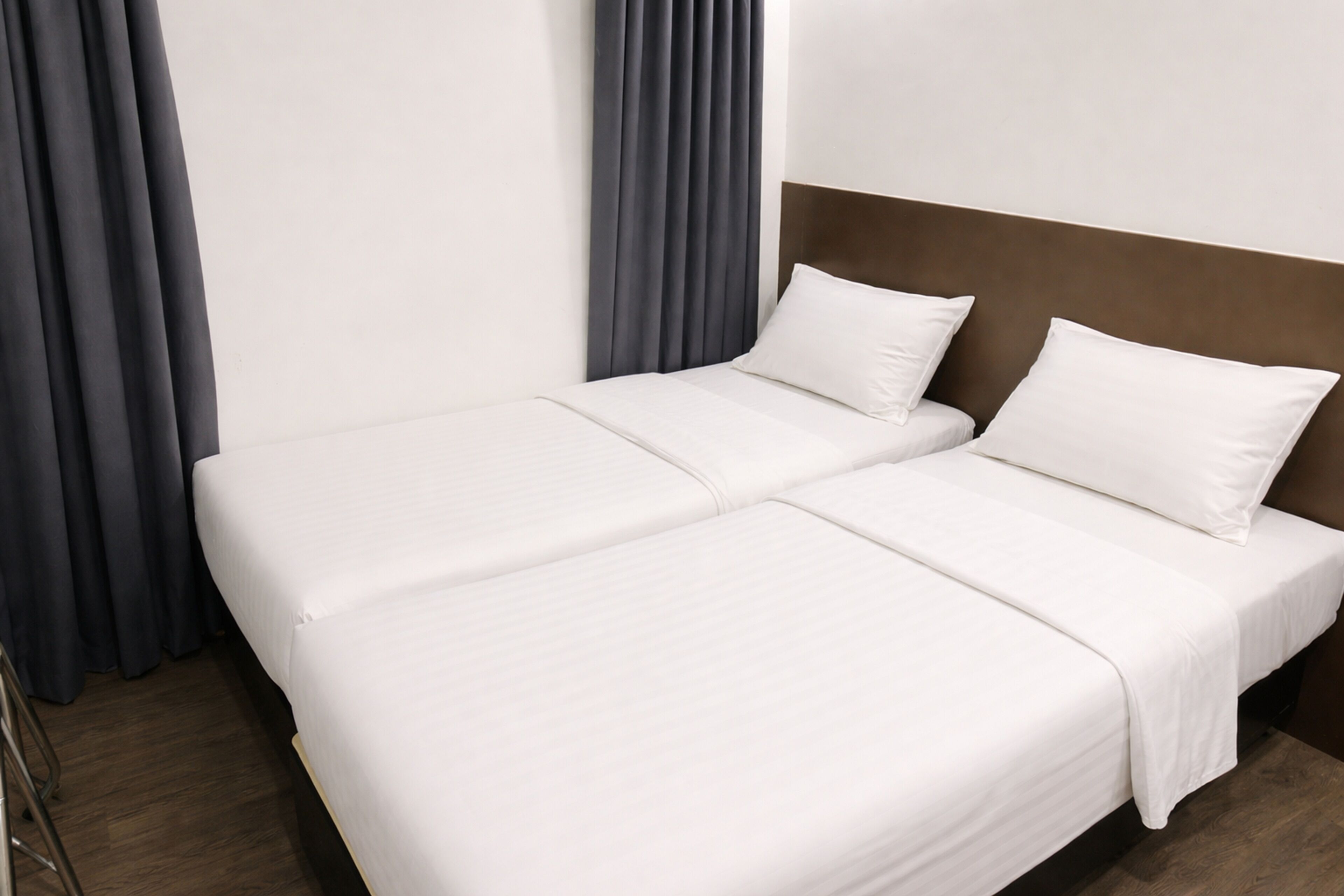 Una cama con dos camas bien hecha, con sábanas blancas y un cabecero de madera, ubicada frente a una pared lisa con una cortina oscura a la izquierda.