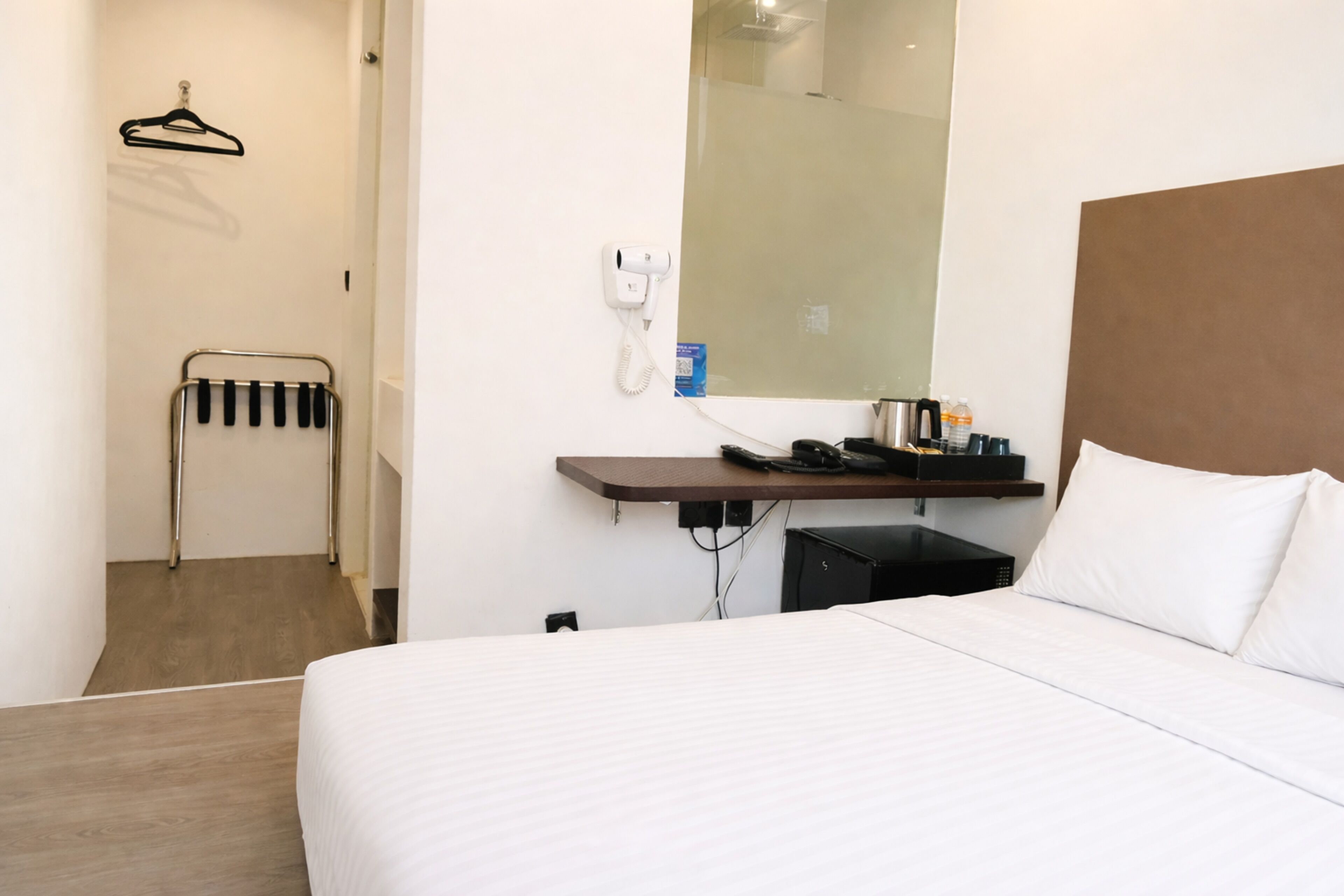 A hotel room with a bed, a bedside table with a phone and toiletries, a towel rack, and a mirror.