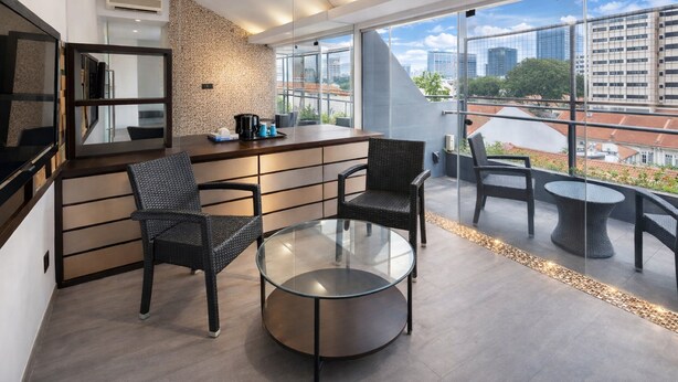 Une chambre d’hôtel moderne avec une table basse en verre, des fauteuils en osier, un coin bar et une vue sur la ville à travers de grandes fenêtres.
