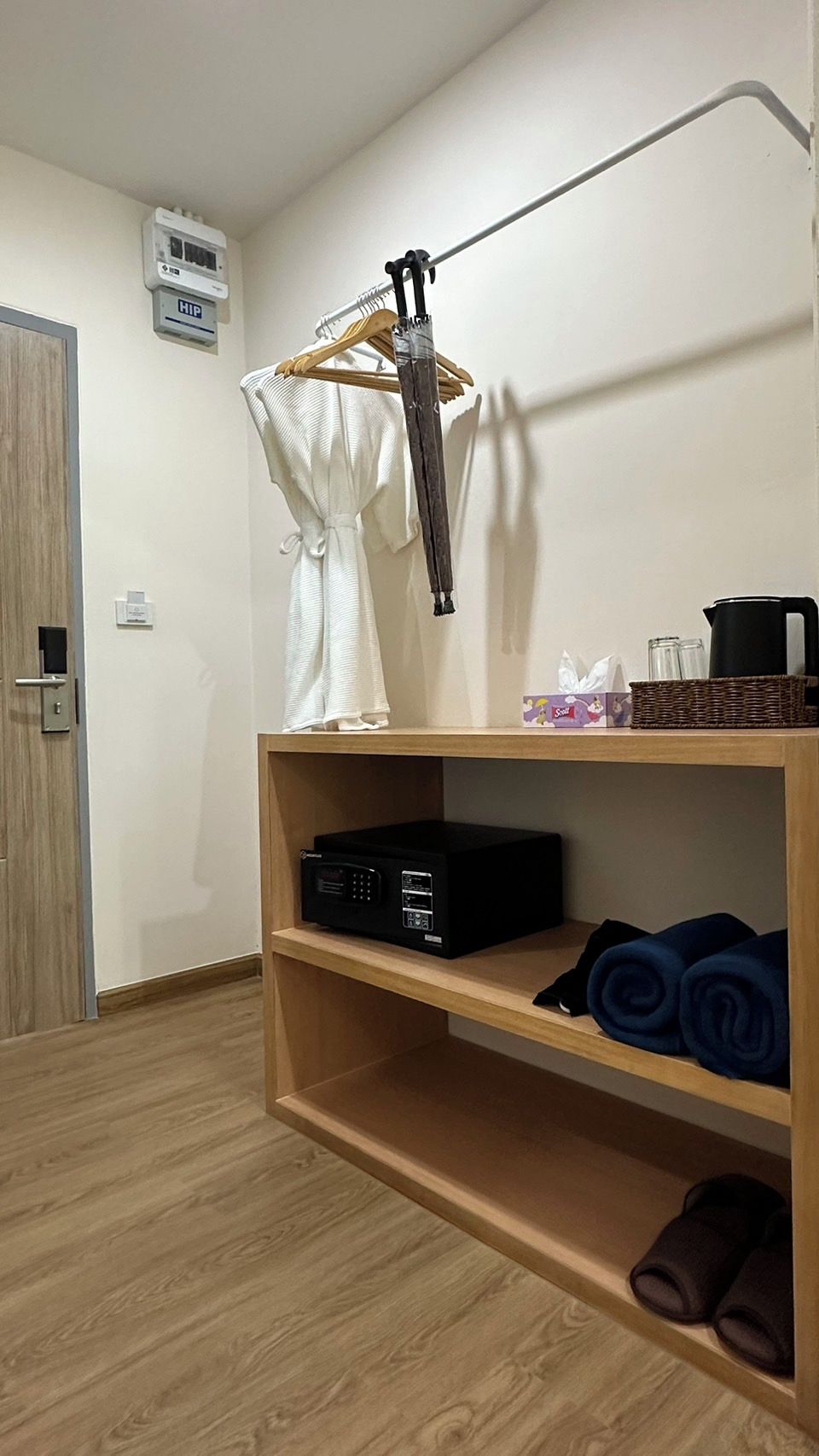 A room with a wooden floor, a wardrobe, a shelf with a radio, rolled towels, a hanging robe, a kettle, and a box of tissues.