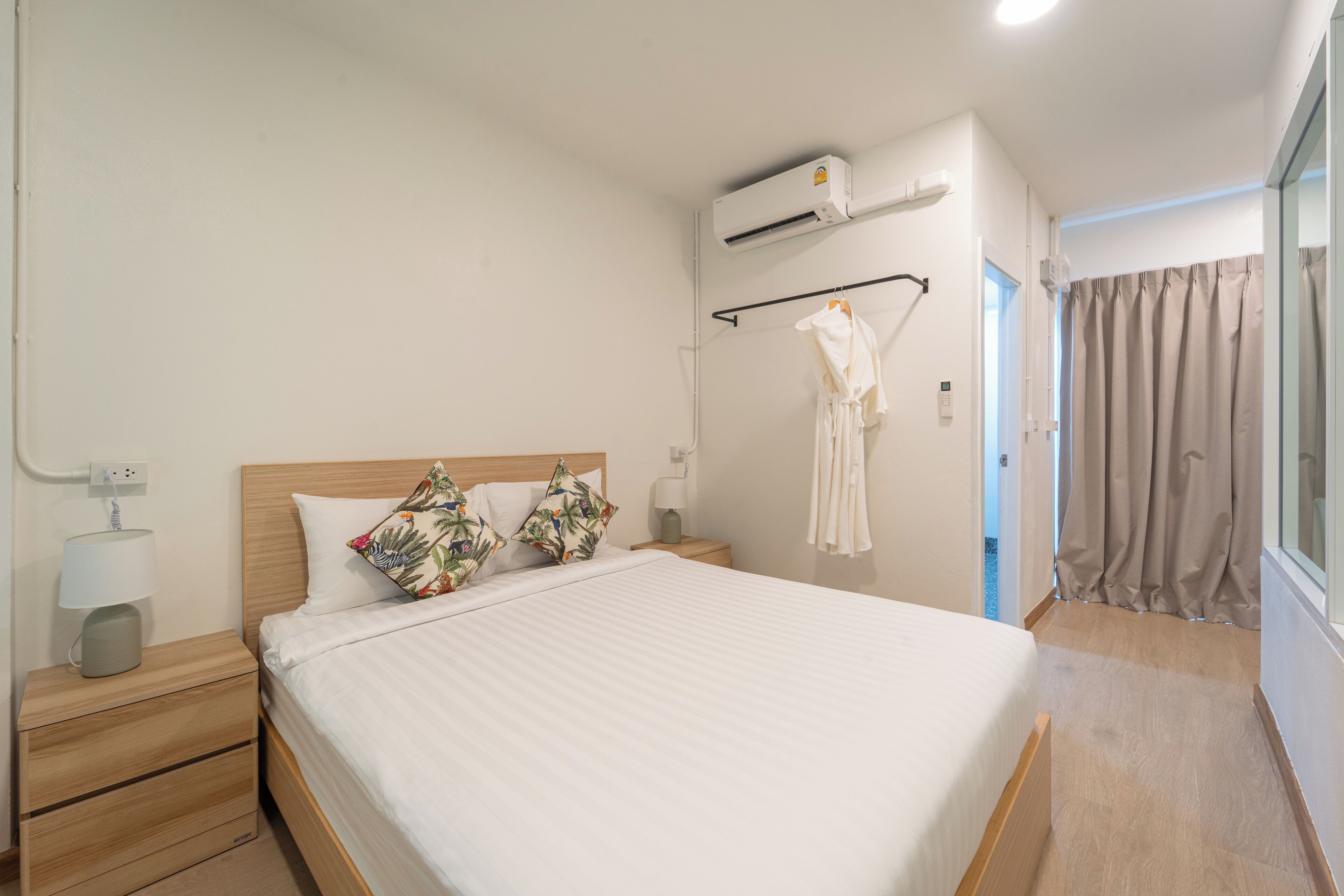 A bedroom with a bed, a nightstand, an air conditioning unit, a robe hanging on a rack, and a view of the outside through a window.