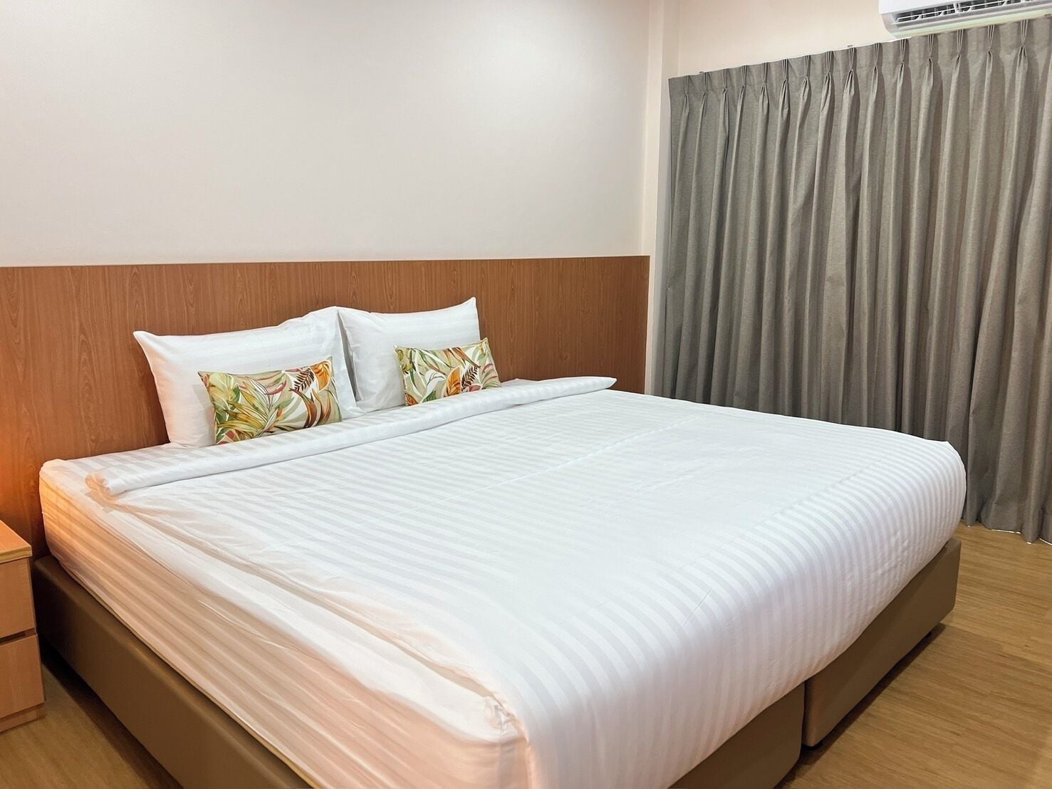 Un lit bien fait, avec du linge de lit blanc et deux coussins décoratifs, une tête de lit en bois, un rideau gris et un plancher en bois.