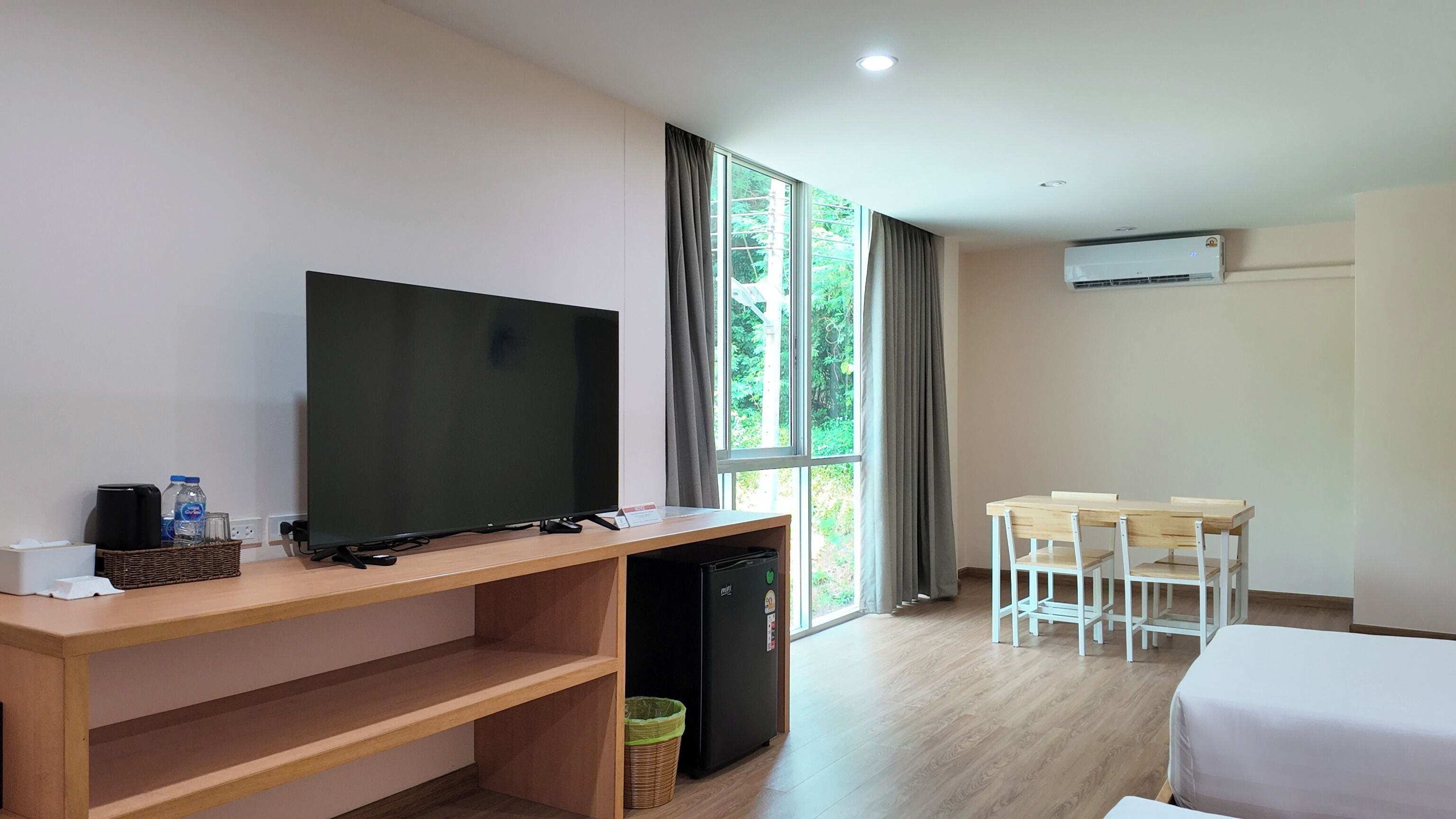 Habitación de hotel con televisión sobre un soporte de madera, un refrigerador pequeño, un escritorio con sillas y ventanas amplias con cortinas.