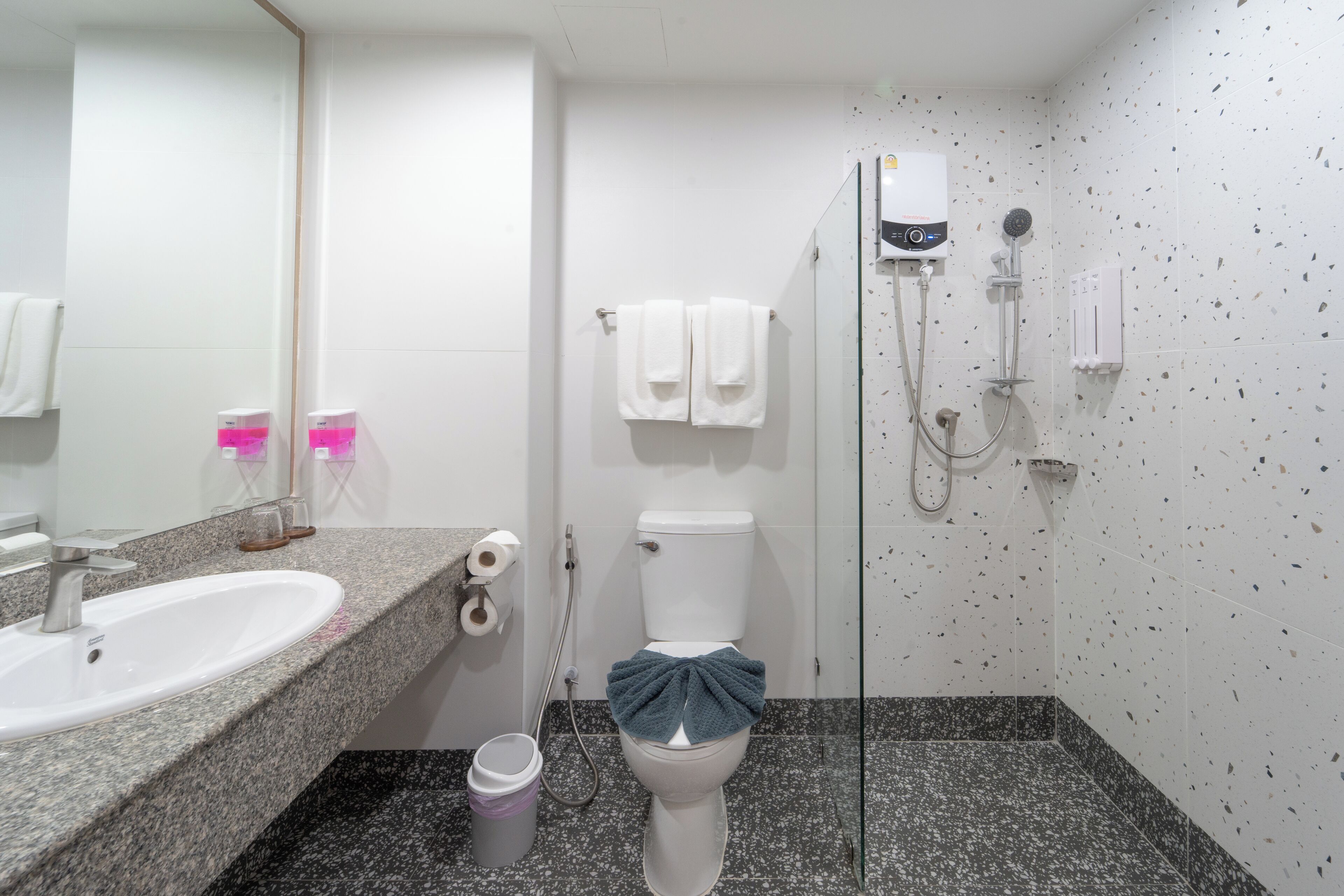 Uma casa de banho com lavatório com bancada de granito, sanita com toalha, chuveiro com duche de mão e dispensador de papel higiénico fixado à parede.