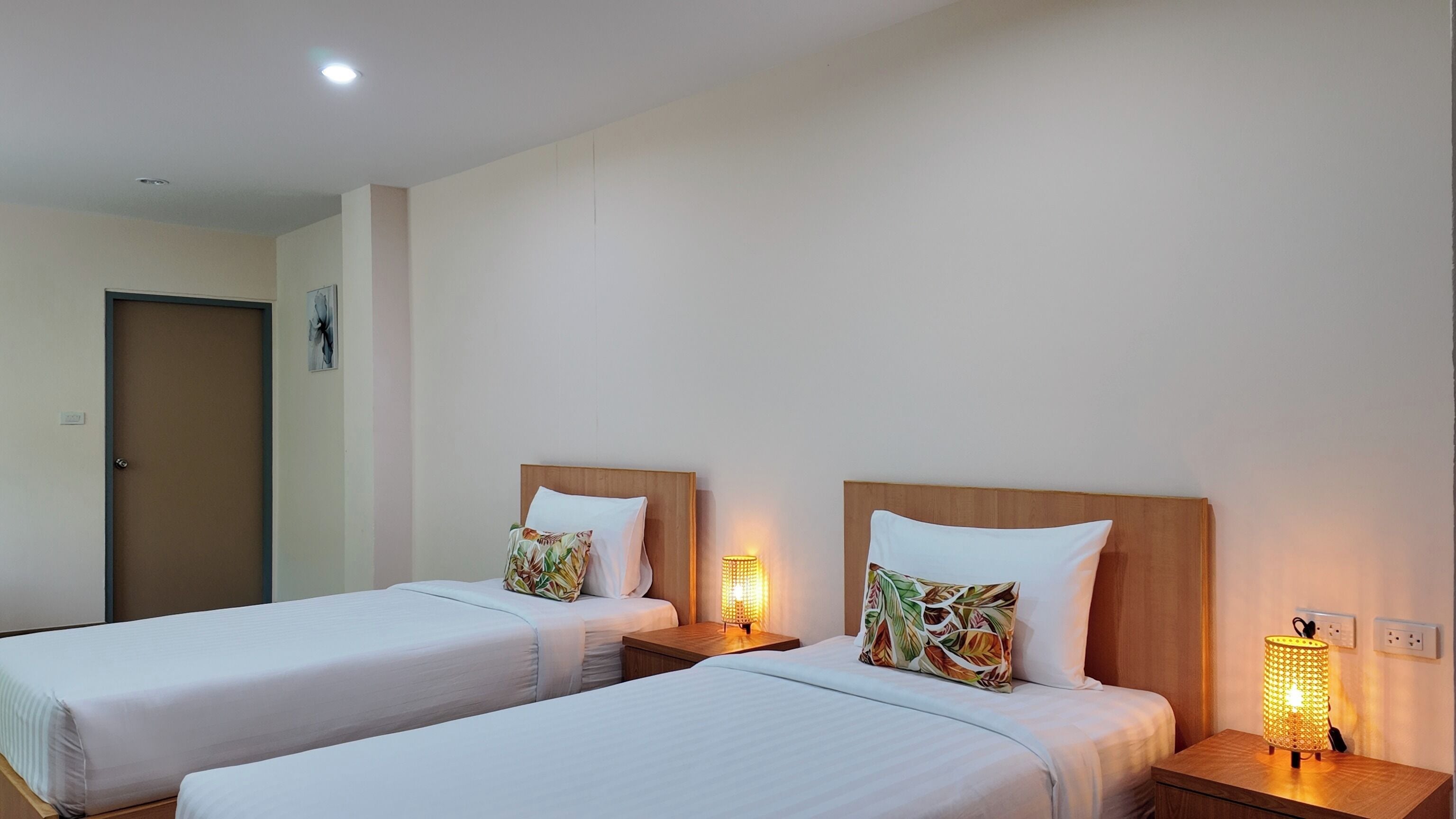 A hotel room with two single beds, each with a wooden headboard, white bedding, and a floral pillow. There are bedside tables with lamps, a door, and a wall-mounted light fixture.