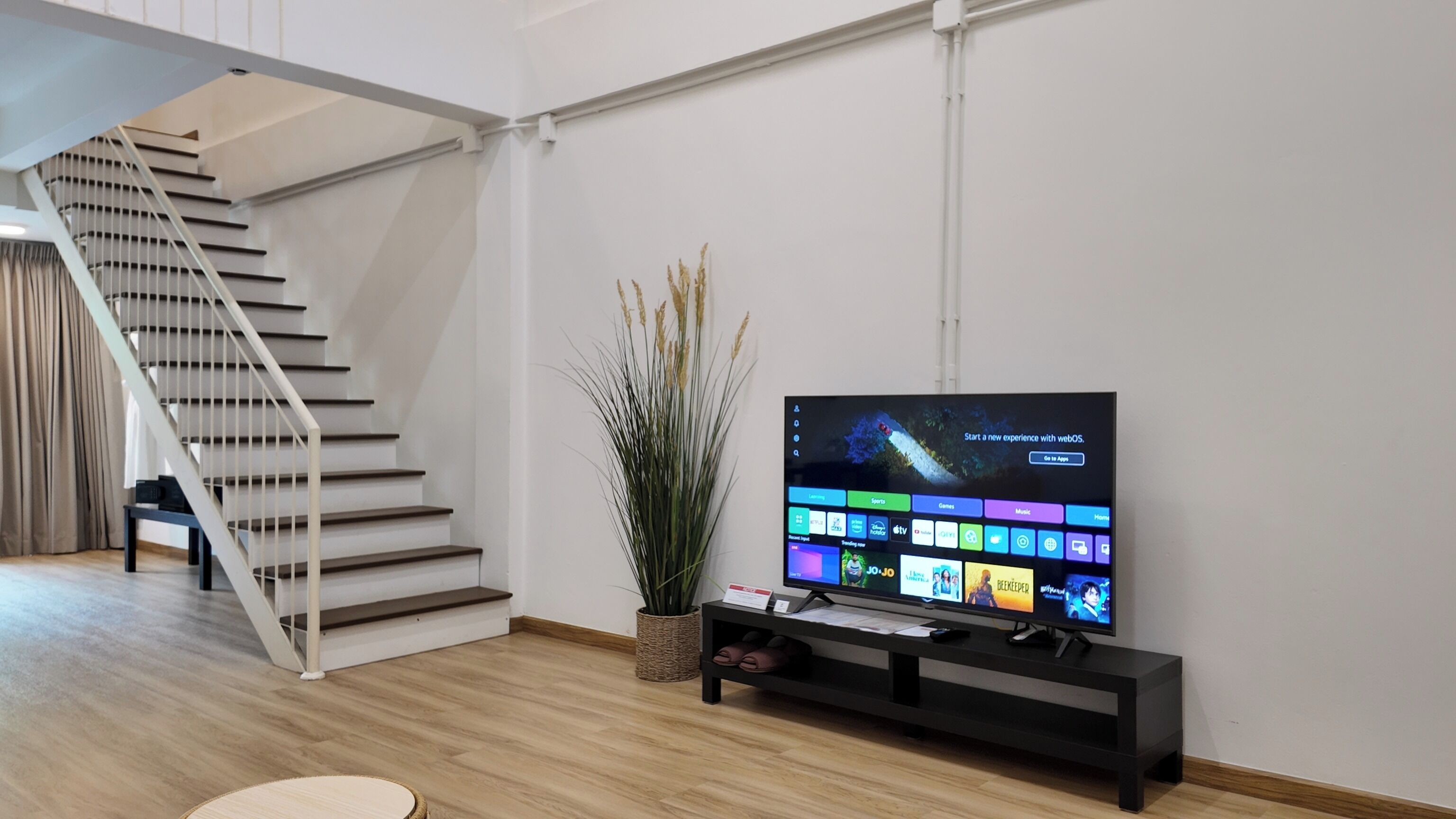 A modern living room with a staircase, a flat-screen TV on a black stand, and a potted plant.