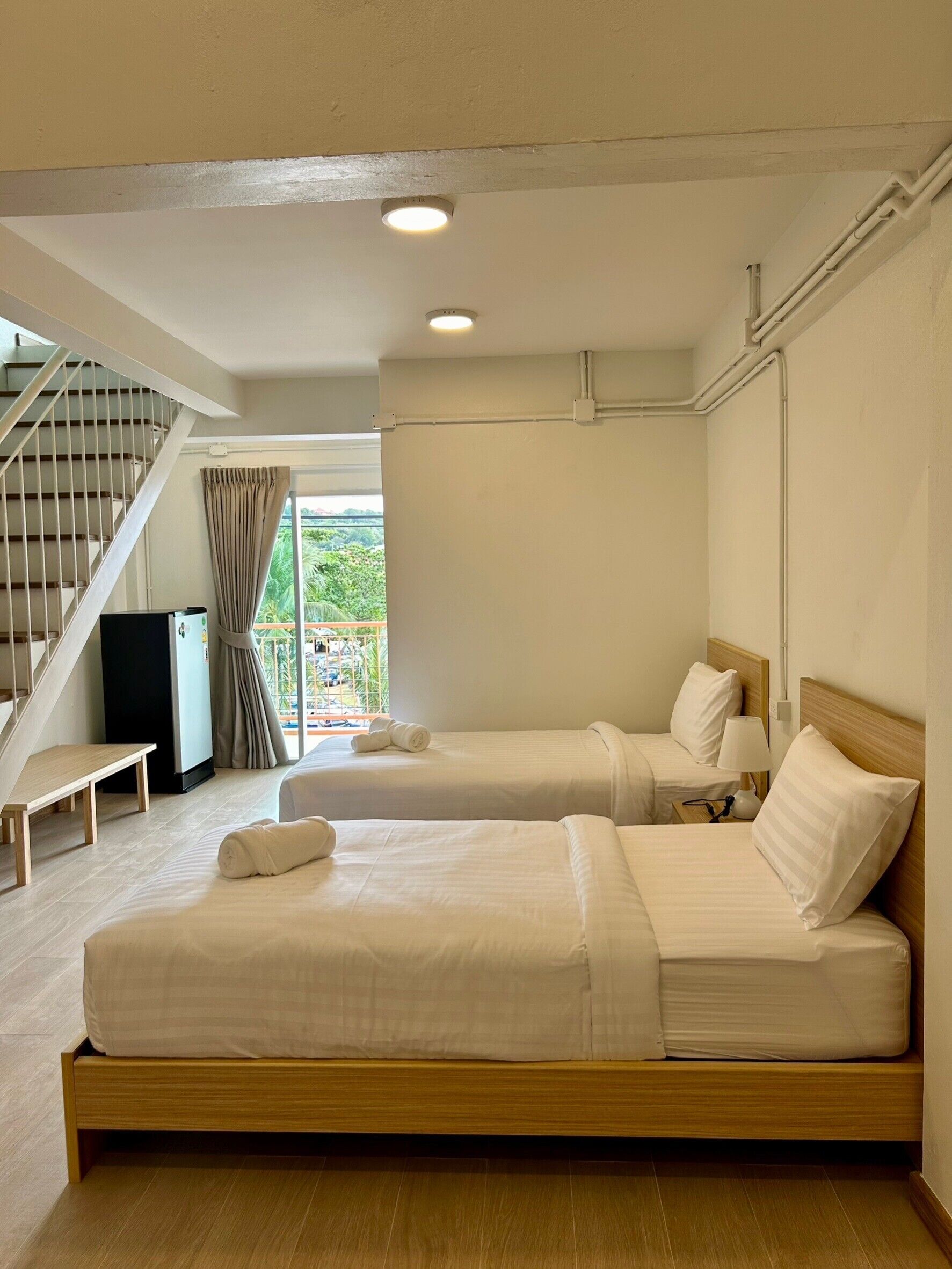 A hotel room with two beds, a wooden bench, a desk, a refrigerator, and a staircase.
