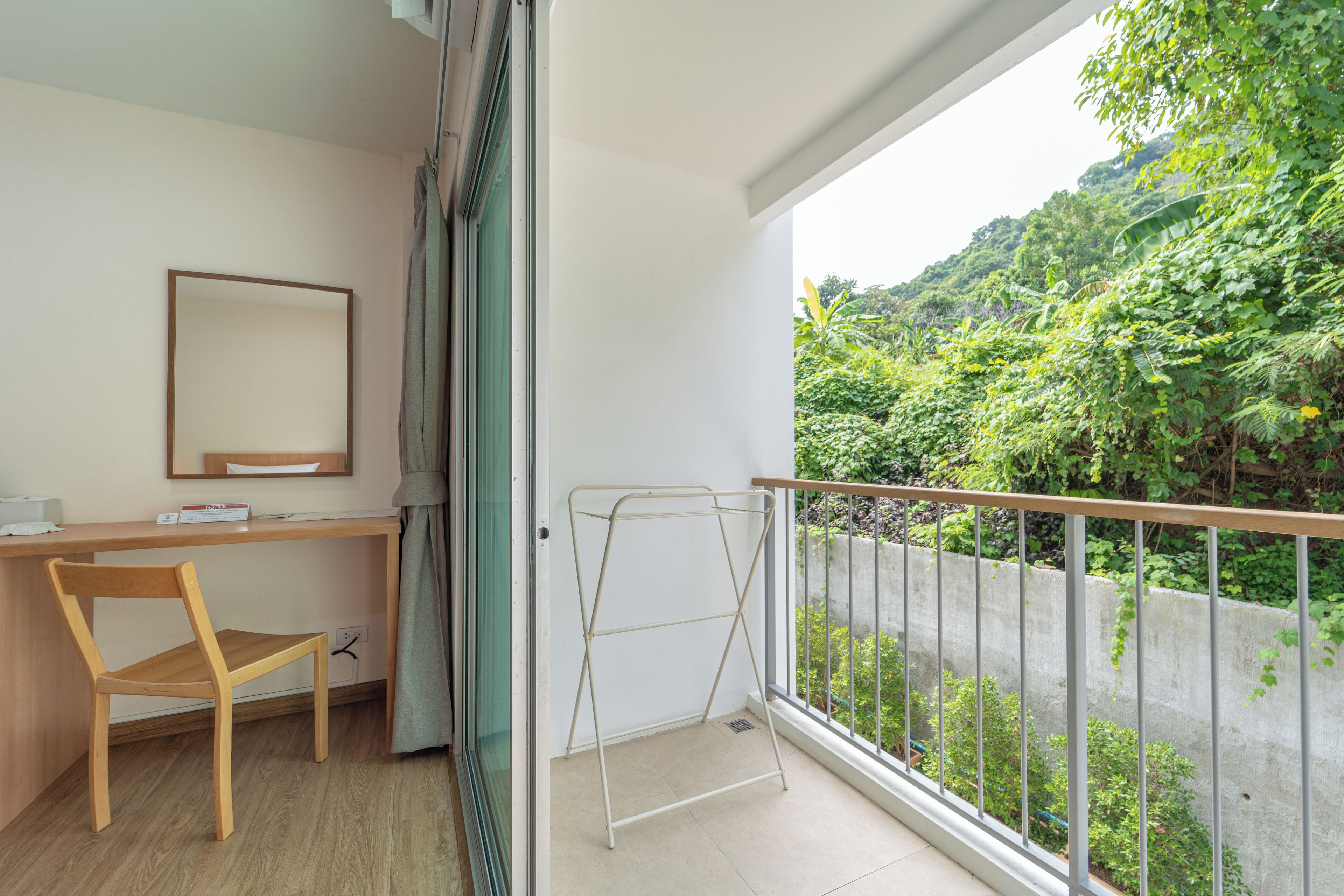 A balcony with a metal railing, a wooden chair, a desk with a mirror, and a view of lush greenery.