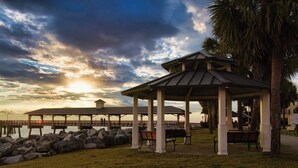 A gazebo with a roof supported by columns, benches, and a view of the water and sunset.