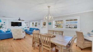 A spacious living room with a dining area, a blue sofa, a television, and a large window with blinds.