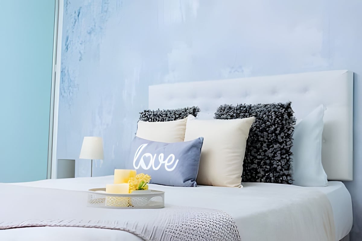 A neatly made bed with a white headboard, a decorative pillow with the word "love," and a bedside table with a lamp and a tray with a candle and flowers.