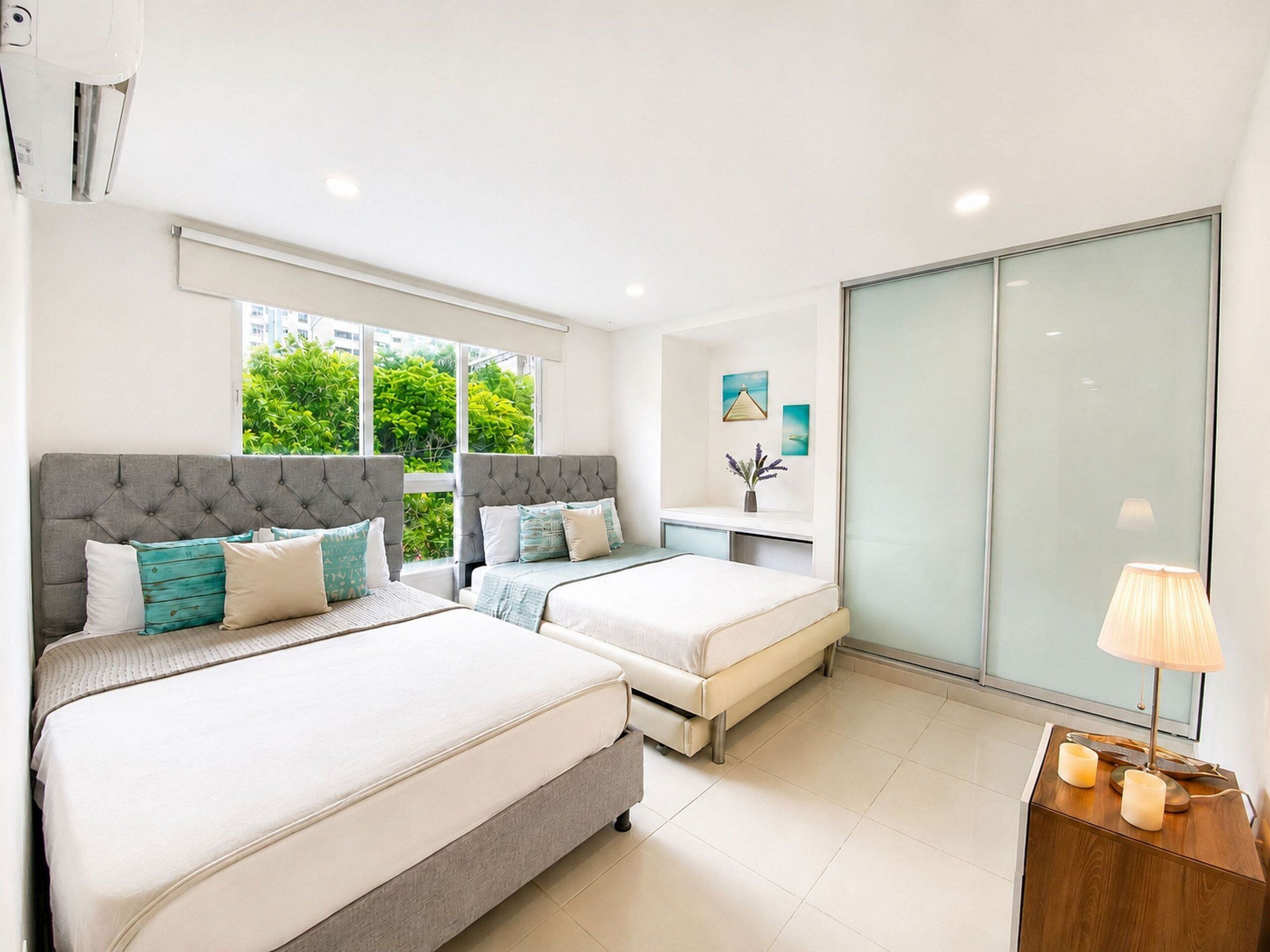A modern hotel room with two beds, a nightstand, a lamp, and a view of greenery outside.