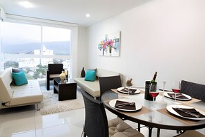 Un salon moderne avec un canapé, une table à manger pour deux personnes et une grande fenêtre donnant sur la ville.