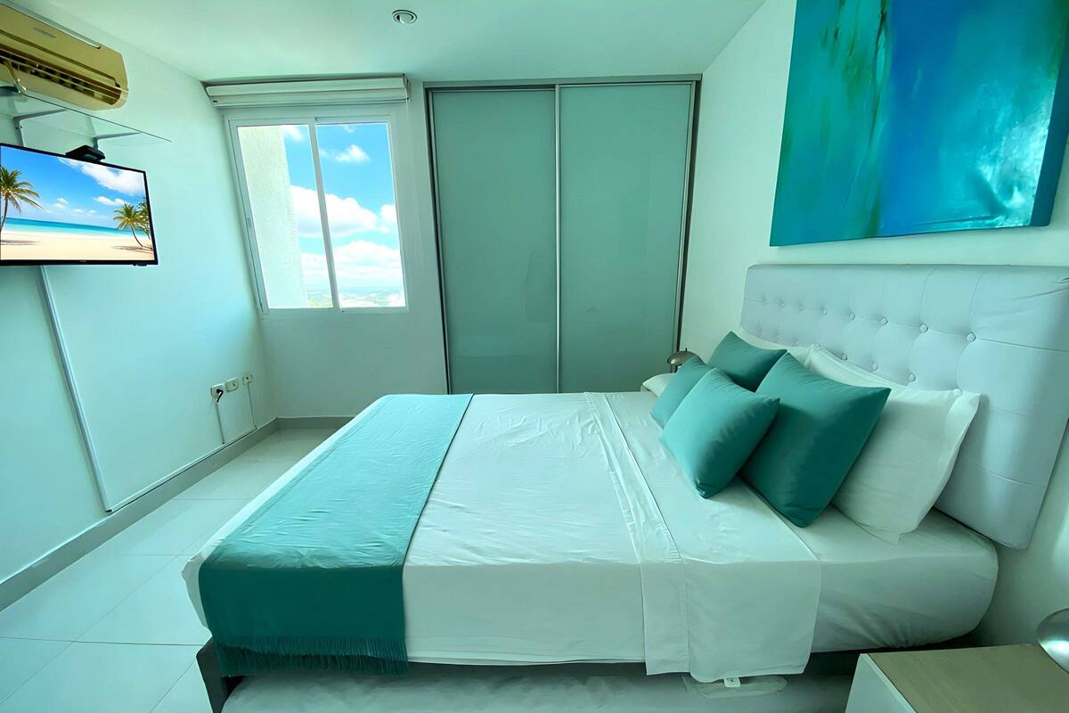 A bedroom with a bed, a television on the wall, a window, and a painting.