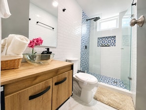 A modern bathroom with a glass shower enclosure, a wooden vanity, a white toilet, and a decorative pink flower. - Apartamentos Neos - Cerca al Mar by SOHO (Santa Marta)
