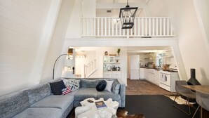 Een loft-achtige leefruimte met een hoge plafond, een trap naar een hoger niveau, een eethoek met een tafel en stoelen, en een keukenhoek.