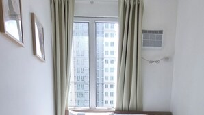 Een hotelkamer met een groot bed, twee kussens, een hoofdeinde, een raam met gordijnen, een wandairconditioner en ingelijste foto's aan de muur.