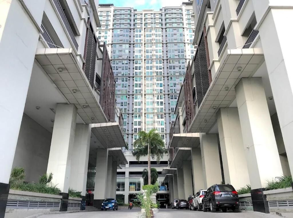 Un edificio residencial de varios pisos con una entrada central, rodeado de automóviles estacionados y una palmera pequeña.