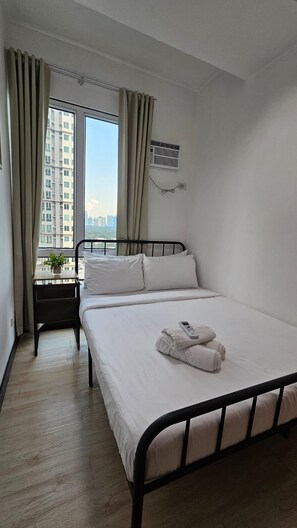 A hotel room with a bed, a bedside table, a window with curtains, and an air conditioning unit.