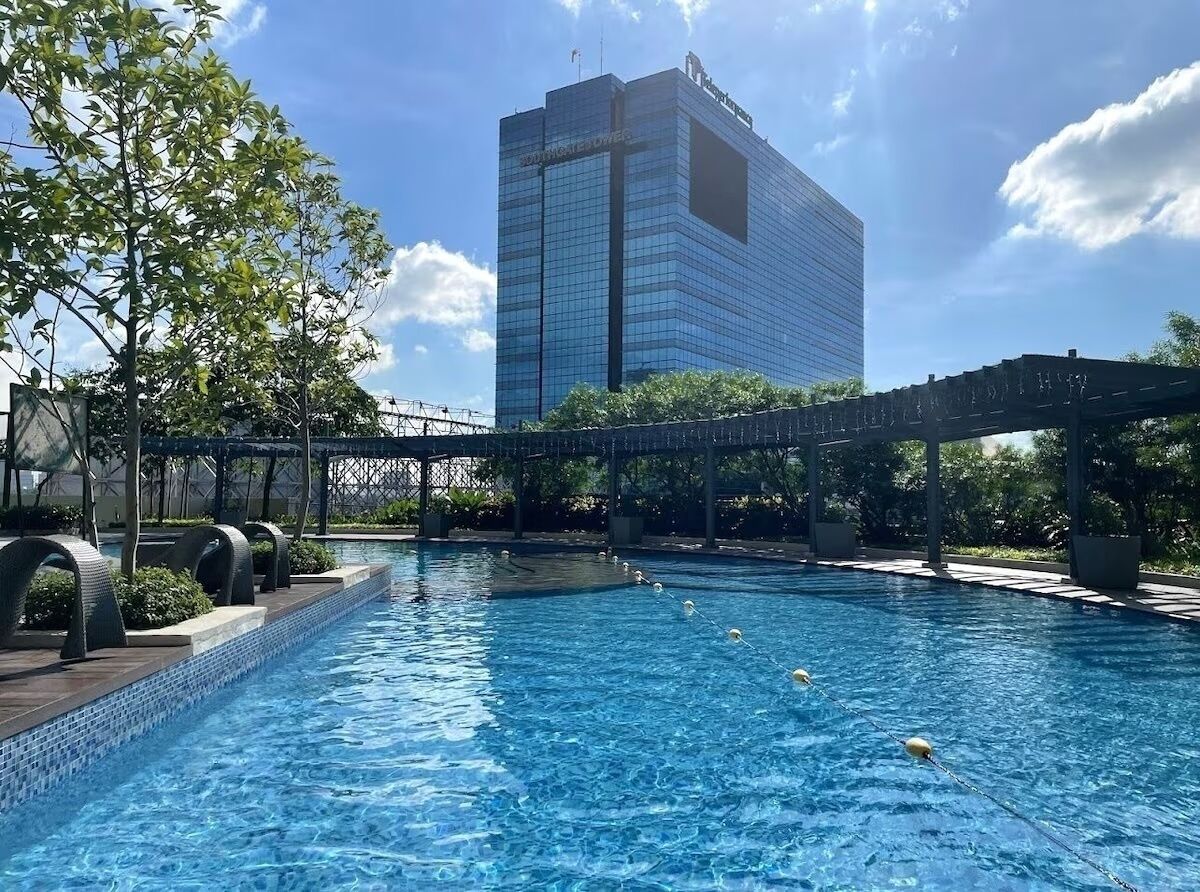 Una piscina con un puente, rodeada de follaje y un edificio moderno al fondo.