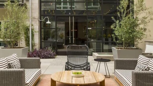 An outdoor seating area with wicker furniture and a round table, surrounded by potted plants.