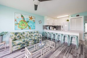 A living room with a sofa, a glass coffee table, and a kitchen area with bar stools. - Paradise Palms 2 Bedroom Condo Ocean Pointe 1206 (Tavernier)