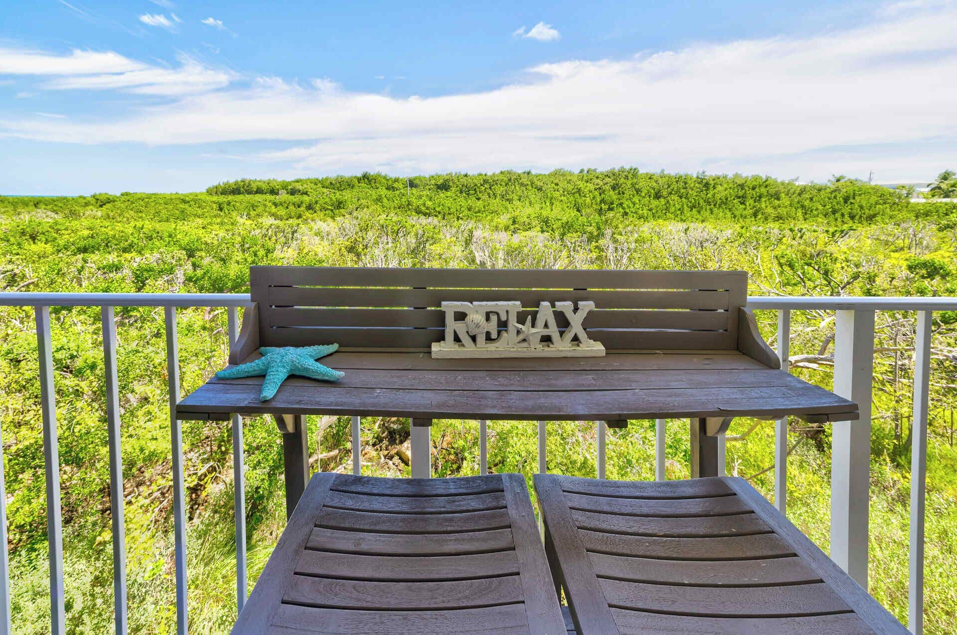 Een houten bank met een zeester en een bord waarop 'RELAX' staat, op een balkon met een leuning, met uitzicht op een weelderig groen landschap.