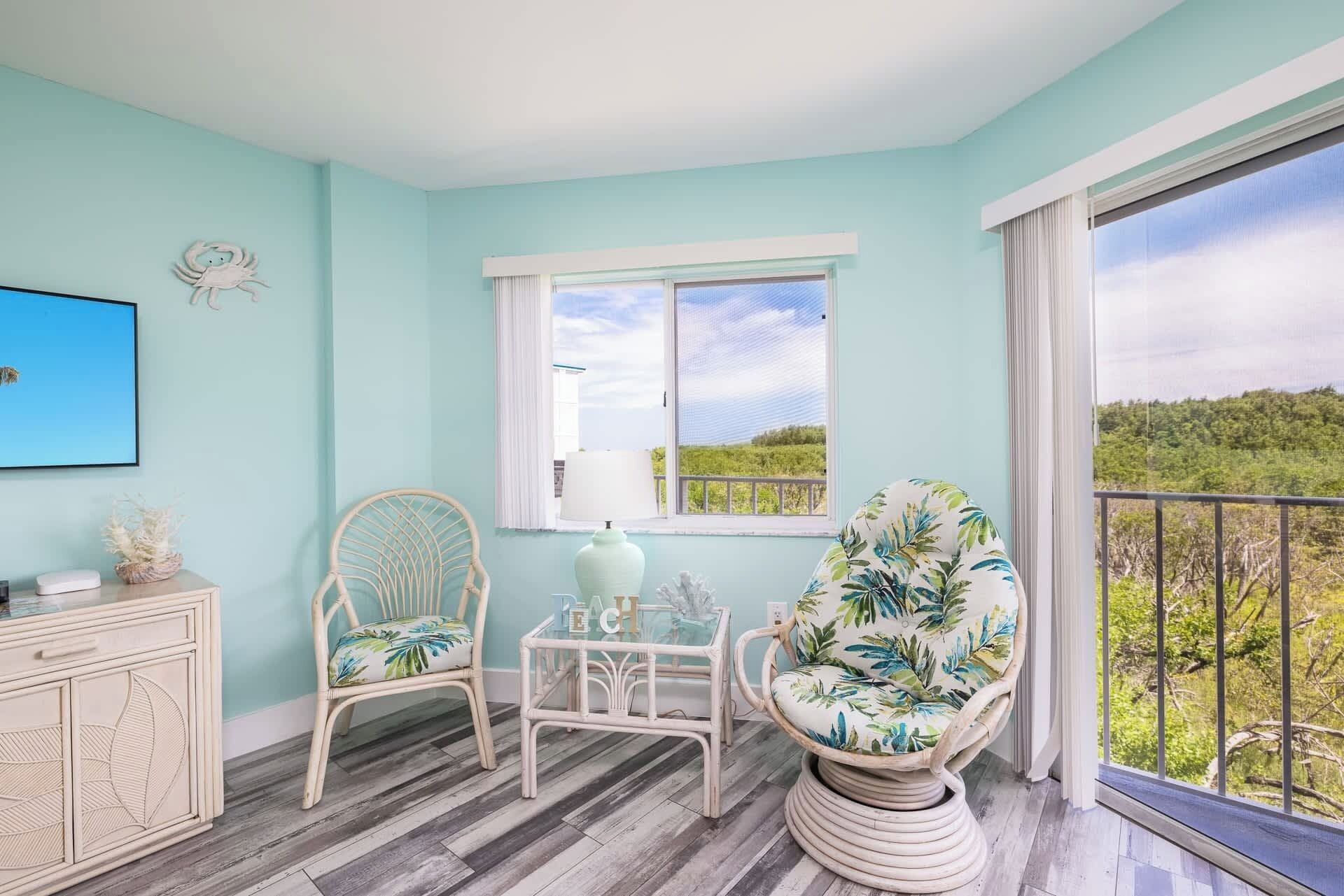 A room with a light blue wall, a wooden floor, a white cabinet, a wicker chair, a glass table, and a view of greenery through a balcony.