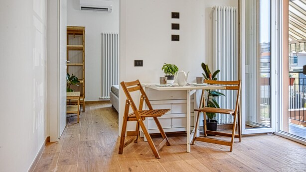 Habitación moderna y bien iluminada con suelo de madera, un escritorio blanco con una silla, un radiador y una ventana con persianas.